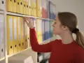 Kobieta w czerwonym swetrze przegląda segregatory, porządkując dokumenty. To część jej zakresu obowiązków.
