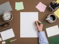 Ręka pisze zielonym ołówkiem na białej kartce. Obok kubek kawy, zeszyt, nożyczki, okulary i koperty. Powstaje ciekawy list motywacyjny.