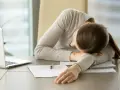 Kobieta z brakiem chęci do pracy odpoczywa na biurku, głowa oparta na ramieniu. Obok laptop i dokumenty.
