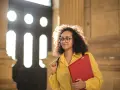 Młoda kobieta w okularach, z kręconymi włosami, trzymająca czerwony segregator, przygotowuje się do ważnego spotkania, być może w celu przedstawienia swojego cv naukowego.