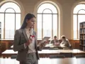 Młoda kobieta z dyplomem w ręku, zastanawia się, czy licencjat to wykształcenie wyższe. W tle biblioteka i studenci.
