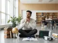 Młody mężczyzna siedzi na podłodze, zastanawiając się nad przyszłymi kierunkami studiów. Wokół niego książki, globus, laptop i model szkieletu.
