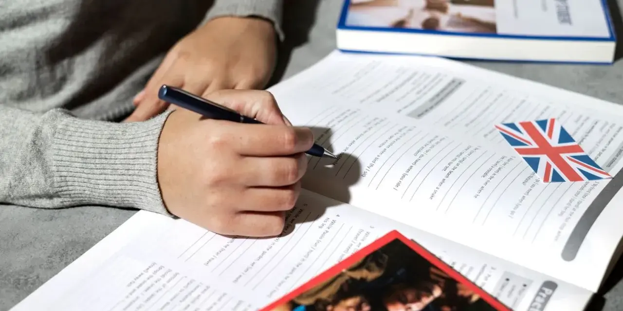 Osoba pisze ćwiczenia z angielskiego, obok leży książka i flaga Wielkiej Brytanii. Klauzula CV po angielsku może być częścią takich materiałów.