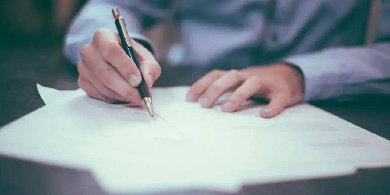 Dłoń pisze długopisem na papierze. Może to być wypowiedzenie umowy, a porzucenie pracy konsekwencje może mieć poważne.