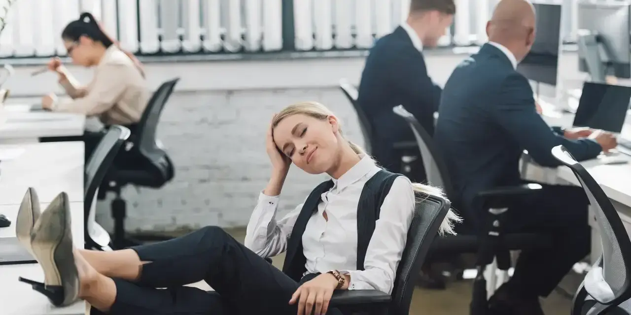Zmęczona pracownica z nogami na biurku, jakby zastanawiała się, czy urlop przechodzi do nowej pracy. W tle inni pracują.