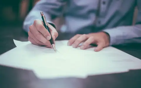 Dłoń pisze długopisem na papierze. Może to być wypowiedzenie umowy, a porzucenie pracy konsekwencje może mieć poważne.