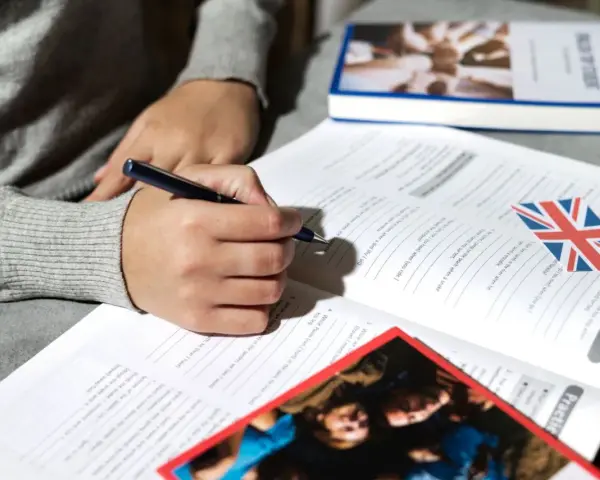 Osoba pisze ćwiczenia z angielskiego, obok leży książka i flaga Wielkiej Brytanii. Klauzula CV po angielsku może być częścią takich materiałów.