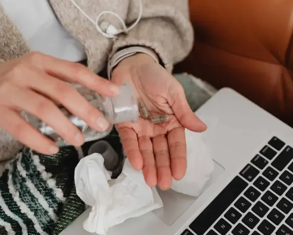 Dłoń z kapsułkami leków, obok laptop i chusteczki. Kto płaci za L4, gdy choroba dopada w domu?