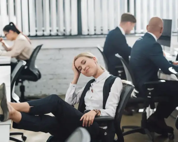 Zmęczona pracownica z nogami na biurku, jakby zastanawiała się, czy urlop przechodzi do nowej pracy. W tle inni pracują.