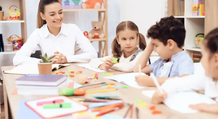 Uśmiechnięta nauczycielka przedszkola z dziećmi przy stoliku, tworząc inspirujący list motywacyjny dla przyszłych pedagogów.