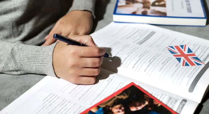 Osoba pisze ćwiczenia z angielskiego, obok leży książka i flaga Wielkiej Brytanii. Klauzula CV po angielsku może być częścią takich materiałów.