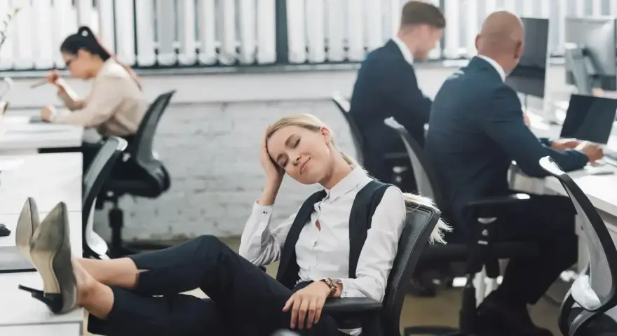 Zmęczona pracownica z nogami na biurku, jakby zastanawiała się, czy urlop przechodzi do nowej pracy. W tle inni pracują.