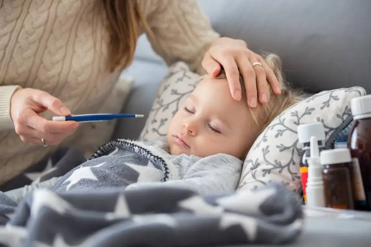Mama sprawdza gorączkę u śpiącego dziecka. Na stoliku leki, a maluch okryty kocykiem z gwiazdkami. L4 na dziecko.