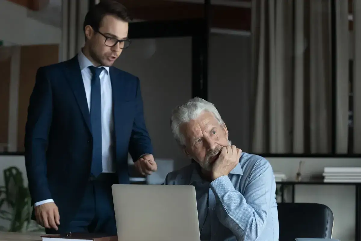 Młodszy mężczyzna w garniturze tłumaczy coś starszemu koledze przy laptopie. To może być pomysł na pracę po 50.
