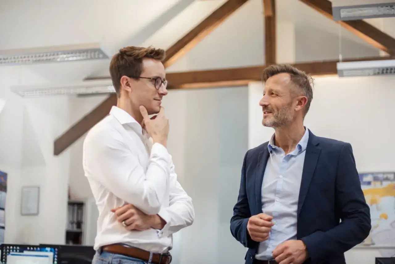 Dw&oacute;ch mężczyzn rozmawia w biurze. Młodszy, w okularach, zamyślony, słucha starszego, kt&oacute;ry z uśmiechem opowiada o możliwościach awansu pionowego.
