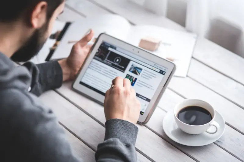 Mężczyzna z brodą przegląda wiadomości na tablecie, popijając kawę. Czyta o umowie na czas nieokreślony.
